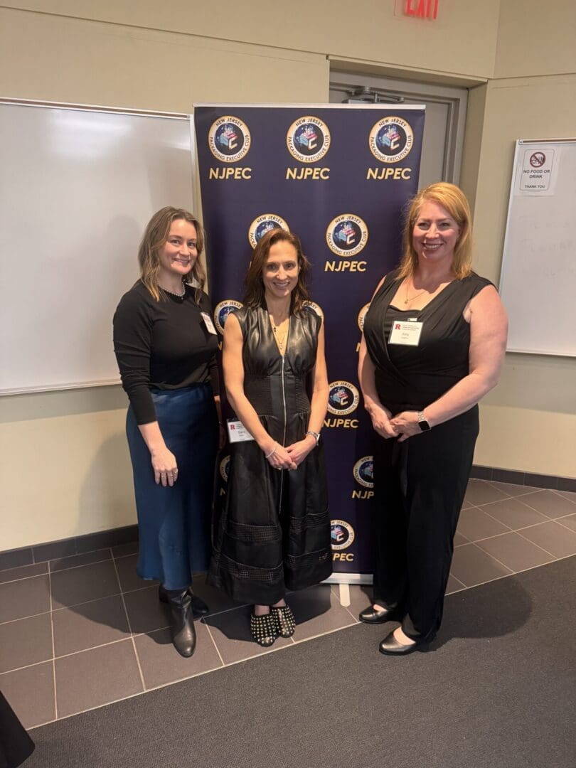 Three women stand smiling in front of an NJPEC backdrop